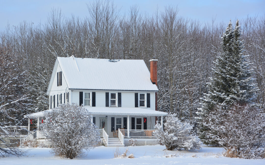 A picturesque white two-story house with a snow-covered roof and a brick chimney, set against a winter backdrop with leafless trees and evergreens. The landscape is blanketed in snow, with a large evergreen tree to the right suggesting the need for winter tree care. A porch adorned with holiday decorations adds a welcoming touch. This image represents 'The Ultimate Guide to Winter Tree Watering - JC Tree Care'.